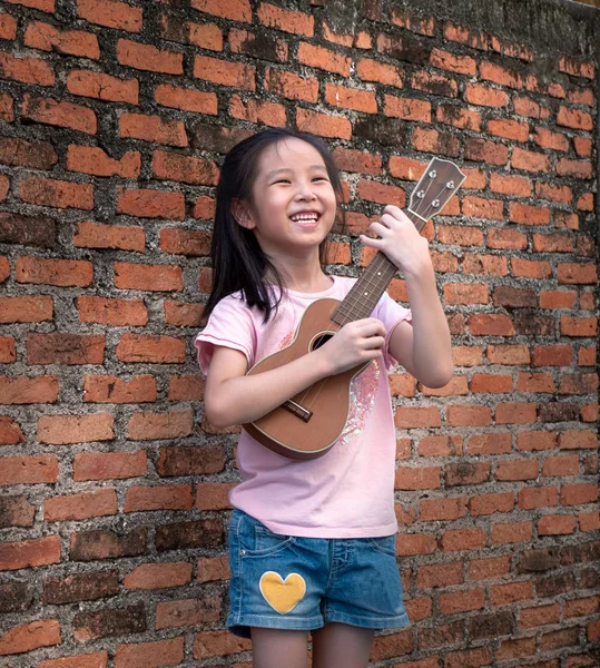 Malé Asijské dítě hrát na ukulele, stará cihlová zeď na pozadí Royalty Free Stock Obrázky