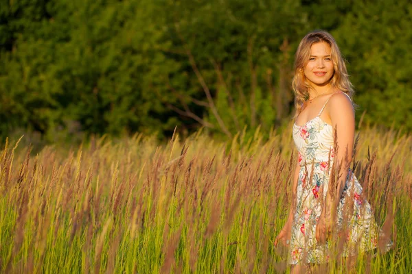 Mulher bonita em pé em um campo, grama verde e flores. Ao ar livre Desfrute da natureza. Menina sorrindo saudável em pé na grama alta — Fotografia de Stock