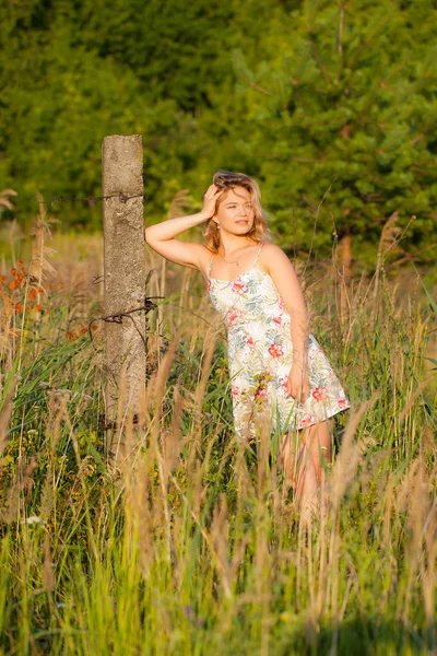 Hermosa joven de pie en un campo cerca de un pilar de hormigón, hierba verde y flores. Al aire libre Disfrute de la naturaleza. Saludable chica sonriente de pie en la hierba alta — Foto de Stock