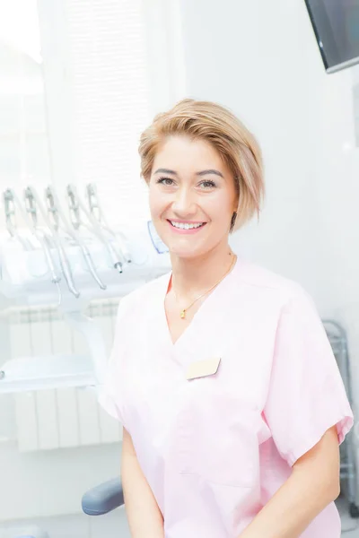 Mujer dentista rubia en una sala de tratamiento, mira a la cámara y sonríe —  Fotos de Stock