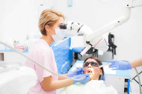Odontóloga joven que trata los conductos radiculares con microscopio en la clínica dental. Paciente joven acostada en la silla del dentista con la boca abierta. Dentista con máscara y guantes — Foto de Stock