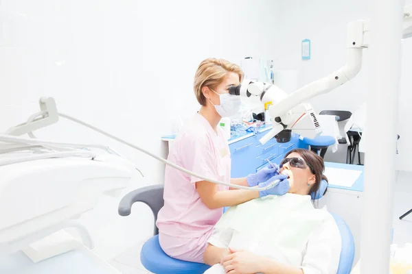 Odontóloga joven que trata los conductos radiculares con microscopio en la clínica dental. Paciente joven acostada en la silla del dentista con la boca abierta. Dentista con máscara y guantes — Foto de Stock