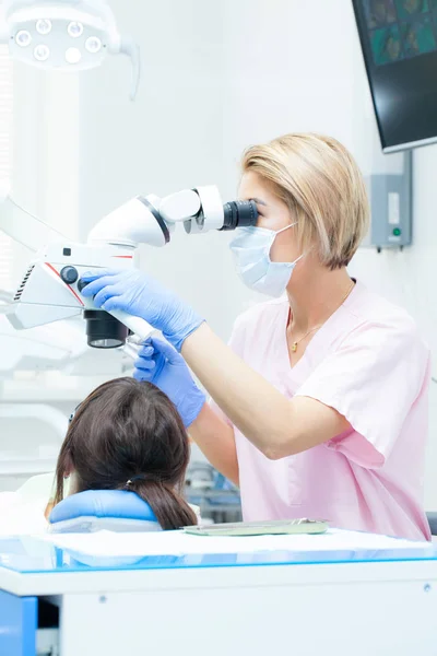 Jeune dentiste qui traite les canaux radiculaires au microscope à la clinique dentaire. Jeune femme patiente allongée sur une chaise de dentiste à bouche ouverte. Dentiste portant masque et gants — Photo