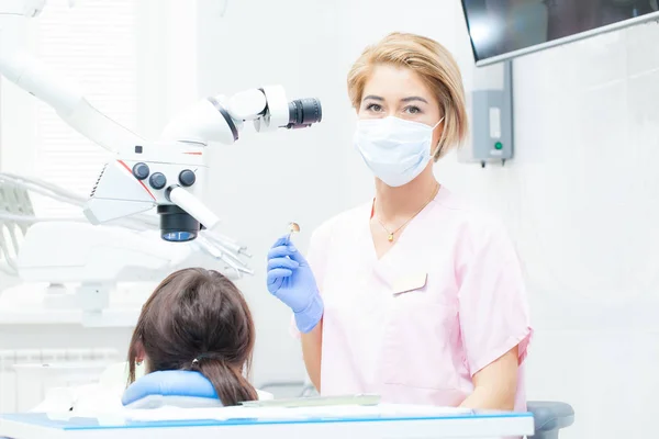 Une jeune dentiste féminine traite les canaux radiculaires au microscope dans une clinique dentaire. Une jeune patiente allongée sur une chaise de dentiste. Un dentiste portant un masque et des gants regarde la caméra — Photo