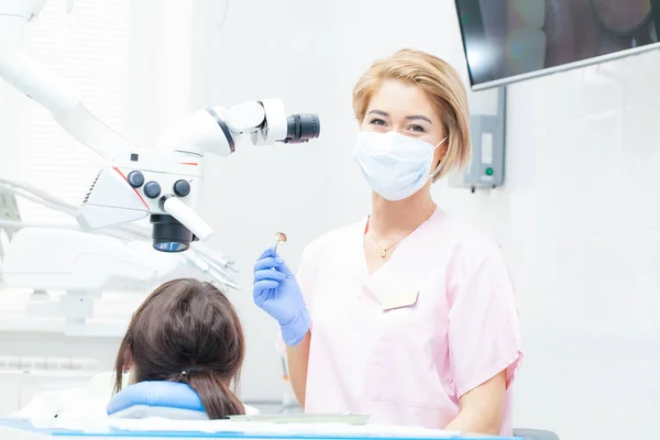 Una giovane dentista sta curando i canali radicolari con un microscopio in una clinica dentistica. Giovane paziente donna sdraiata sulla sedia di un dentista. Un dentista che indossa una maschera e guanti guarda la macchina fotografica — Foto Stock