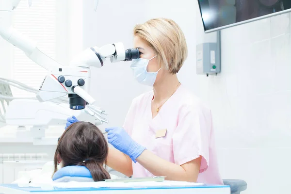 Jeune dentiste qui traite les canaux radiculaires au microscope à la clinique dentaire. Jeune femme patiente allongée sur une chaise de dentiste à bouche ouverte. Dentiste portant masque et gants — Photo
