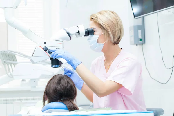 Giovane dentista femminile che cura i canali radicolari utilizzando il microscopio presso la clinica dentale. Giovane paziente donna sdraiata sulla sedia del dentista a bocca aperta. Dentista con maschera e guanti — Foto Stock