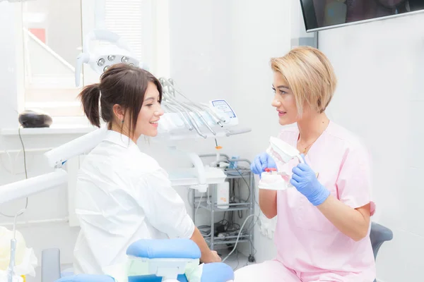 Een vrouwelijke tandarts legt aan een vrouwelijke patiënt met het proces van het tandenpoetsen op een mock-up. Beide kijken naar de camera — Stockfoto