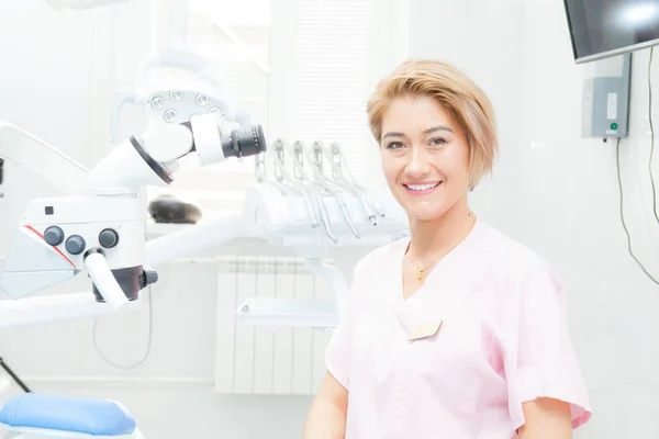 Fille dentiste blonde dans une salle de traitement, regarde la caméra, près d'un microscope médical — Photo