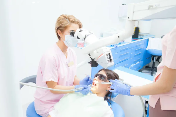 Odontóloga joven que trata los conductos radiculares con microscopio en la clínica dental. Paciente joven acostada en la silla del dentista con la boca abierta. Dentista con máscara y guantes — Foto de Stock