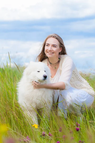 Młoda kobieta siedzi z duży biały pies kudłaty na tle rzeki w zielonej trawie — Zdjęcie stockowe
