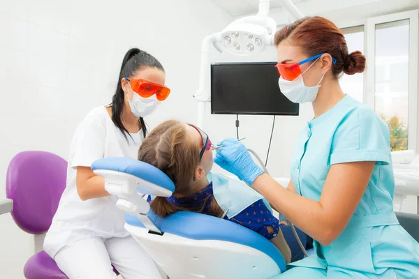 Una mujer joven es dentista ortodoncista y una asistente femenina trata los dientes de una paciente acostada en una silla dental en una clínica dental. Una paciente joven —  Fotos de Stock