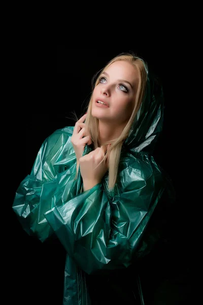 Retrato de uma jovem mulher loira bonita em uma capa de chuva verde olhando para cima, um capuz é colocado em sua cabeça. Tiro em um estúdio de fotos em um fundo preto — Fotografia de Stock