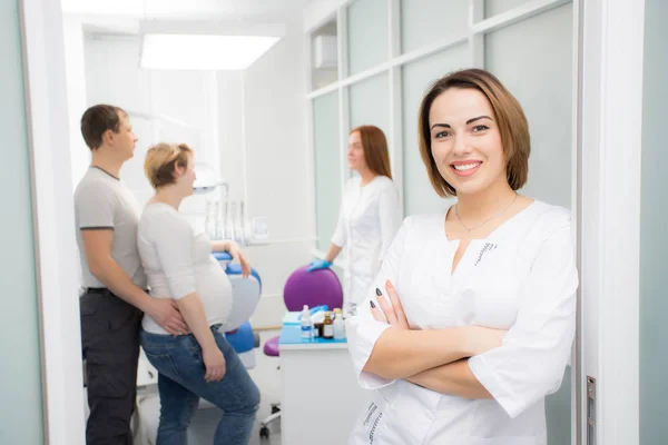 Mooie jongedame arts staat tegen de achtergrond van de patiënten in de kliniek. Patiënten gehuwd paar, vrouw zwanger — Stockfoto