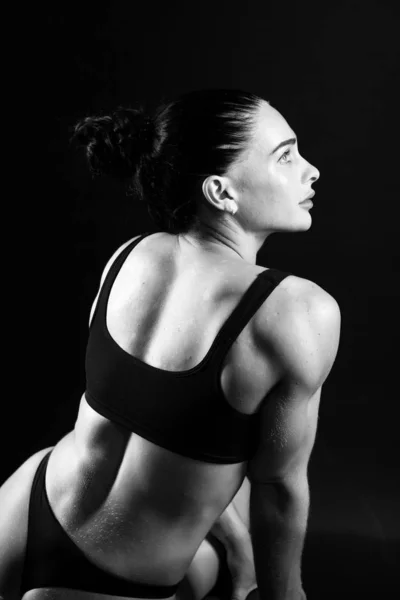 Монохромное фото молодой атлетичной женщины спорта, построенной в купальнике. Тематический спорт и бодибилдинг. Женщина на черном фоне, сидящая спиной к полному росту — стоковое фото