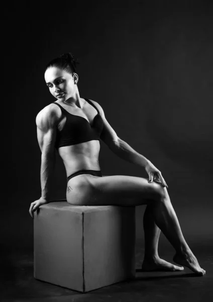 Monochrome photo of a young athletic woman sports build in a swimsuit. Theme sports and bodybuilding. Woman on a black background sitting back to full growth — Stock Photo, Image