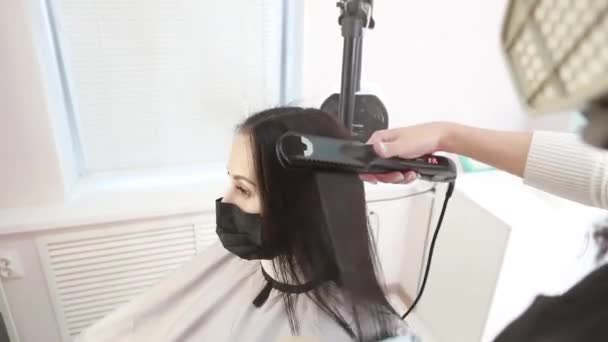 Mujer hermosa joven en el procedimiento de tratamiento del cabello de queratina. Joven peluquera en máscara protectora trabaja con el pelo en el salón — Vídeo de stock