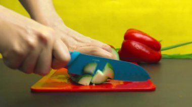 Bir kız kırmızı plastik kesme tahtasında mavi bir bıçak ile sarı bir arka planda bir üçgen şeklinde bir salatalık kesme. Siyah mutfak masası, sebzeler, yeşillikler. 