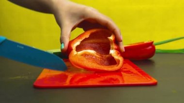 Kız kırmızı plastik kesme tahtası üzerinde mavi bir bıçak ile tatlı biber keser. Sarı arka plan, siyah mutfak masası, sebze, yeşillik. Close.