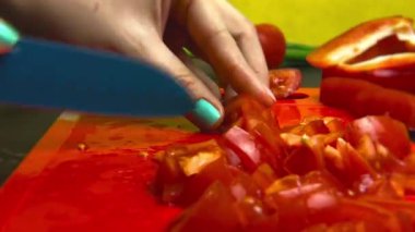 Kız ince bir kırmızı plastik kesme tahtası üzerinde mavi bir bıçak ile bir domates keser. Sarı arka plan, siyah mutfak masası, sebze, yeşillik. Close.