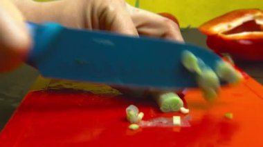 Kız kırmızı plastik kesme tahtası üzerinde mavi bir bıçak ile yeşil soğan tüyleri keser. Sarı arka plan, siyah mutfak masası, sebzeler, yakın çekim.