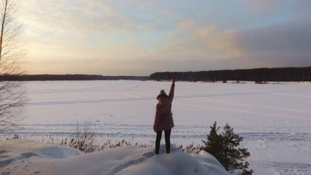 Quadcopter 夕暮れ時 冬の凍った湖の上飛ぶ 崖の上に立っているダウン ジャケットの長いピンクの女の子とコーヒーのプラスチック製のコップをピックアップします 雪の中でスノーモービルのトレイル 美しい空 — ストック動画