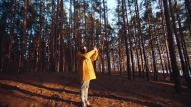 Sarı ceketli bir kadın baharda bir tepenin kenarındaki çam ormanında altın saatindeki parlak güneşi fotoğraflıyor. Doğa, seyahat.