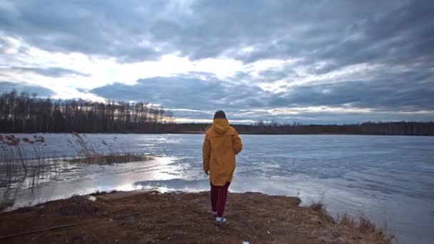 黄色のジャケットを着た女性が森の中の凍った湖のほとりに立って 携帯電話で曇り空の写真を撮る 春の旅行 — ストック動画