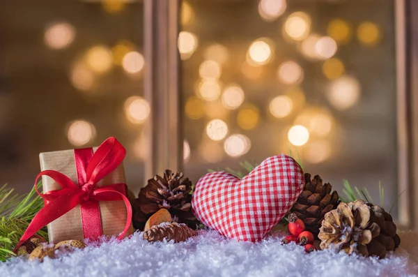 Christmas present and red heart with natural ornaments on snow and blurred light background