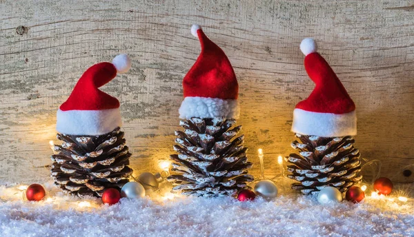 Christmas background with three pine cones decorated with santa caps, red and white baubles on snow with lights on wood background