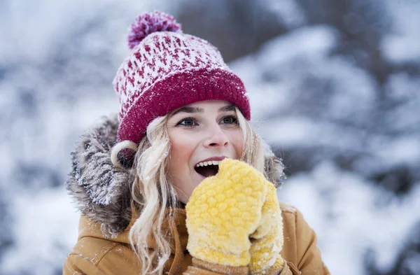 Very beautiful bright young girl in a red hat and yellow mittens on the background of a winter park in the snow. Winter holidays concept. Throw and blow snow