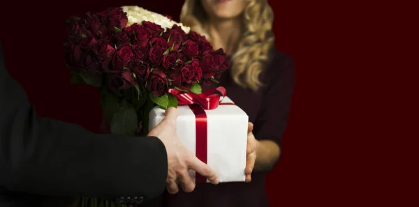 A young man gives a gift a white box with a red bow and flowers to a girl on an isolated black background. Valentine day concept. Lovers day.