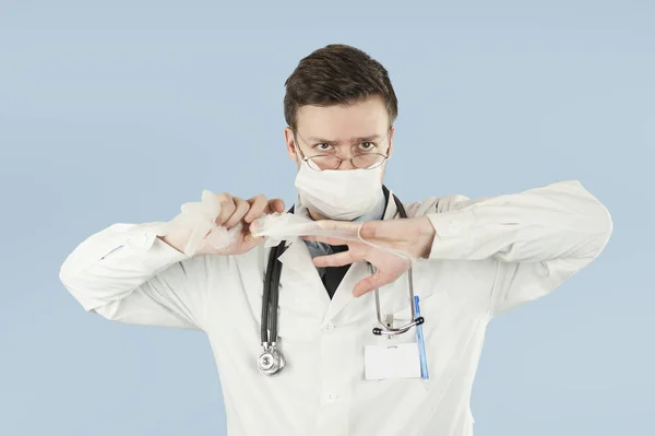 A young fellow doctor student removes disposable hygiene gloves by tearing them against a blue isolated background.