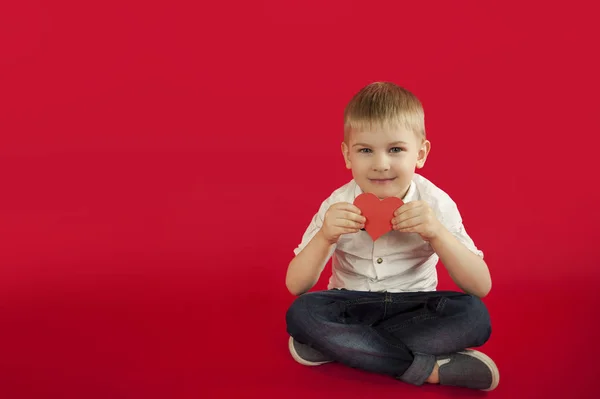 for holidays, lovers day and mothers day, baby boy sits on a red background with a red heart in his hands. The concept is to give gifts and love