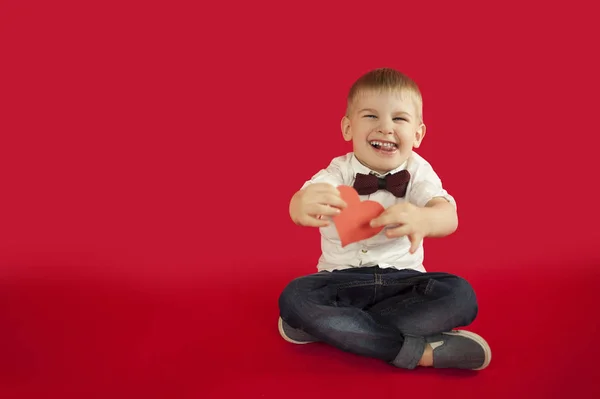 for holidays, lovers day and mothers day, baby boy sits on a red background with a red heart in his hands. The concept is to give gifts and love