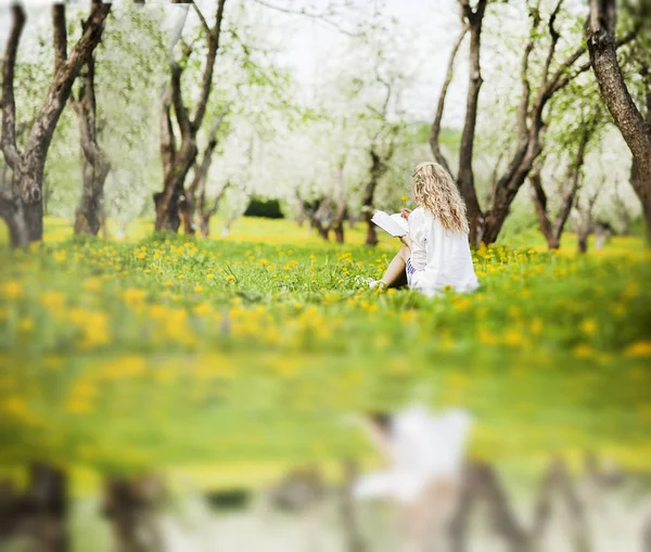 Mladá krásná blondýnka s krásnými vlasy sedí v parku s knihou a květinou v rukou — Stock fotografie
