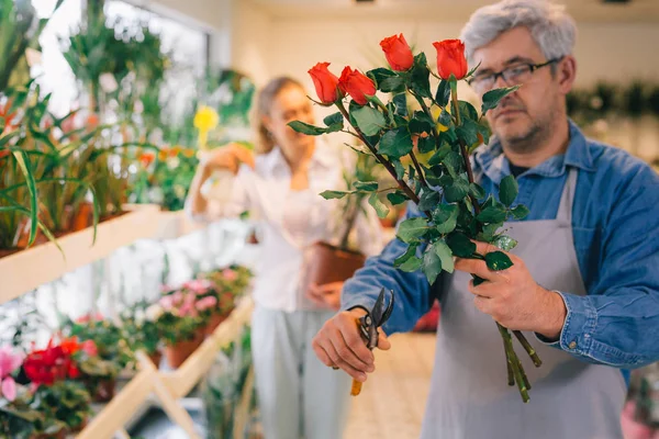 Mature florist trimming roses, woman worker in blurred background.