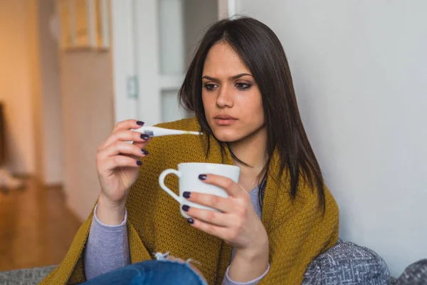 生病的女人看着温度计 而坐在沙发和喝茶回家 — 图库照片