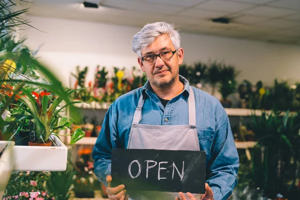 Mature florist man holding open sign in flower shop.