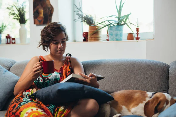 woman relaxed in sofa with dog reading book and drinking coffee
