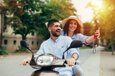 Genç mutlu çift dışarıda motosikletle geziyor ve parlak güneş ışığıyla fotoğraf çekiyorlar.