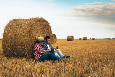 Father and son sitting beside bales using tablet on filed clipart