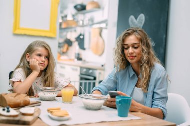 Anne ve kızı evde kahvaltı yapıyor, kız sıkıcı, kadın akıllı telefon kullanıyor..