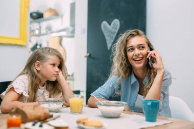 Anne ve kızı evde kahvaltı yapıyor, kız sıkıcı, kadın akıllı telefon kullanıyor..