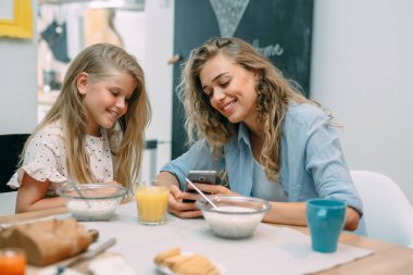 Anne ve kızı kahvaltıda akıllı telefon kullanıyor.