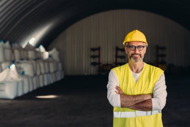 Kasklı işçi depoda çapraz kollar arasında duruyor.