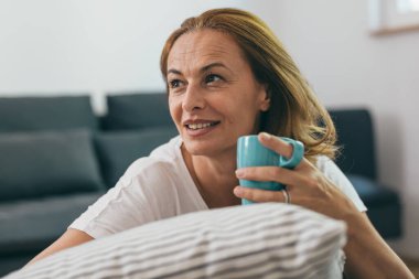Evinde elinde bir fincan kahveyle yastığa yaslanmış güzel olgun bir kadının portresi.