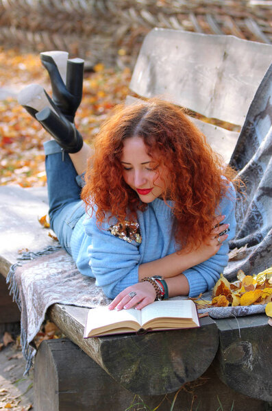 Woman with red hair in the autumn park. lying on a bench with a veil and reading a book. Autumn background