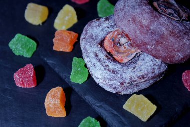 karanlık bir arka plan üzerine bir taş yemek üzerine pudra şekeri yalanlarla Trabzon hurması kurutulmuş. Yakında çok renkli şekerlenmiş meyve parçaları dağılmış