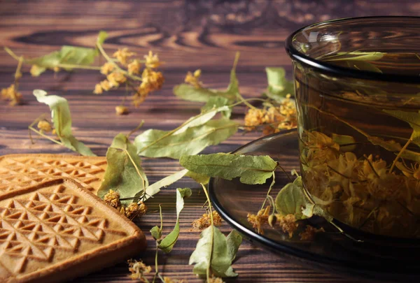 A dark cup of health tea, linden flowers and squares of cookies are on the table.